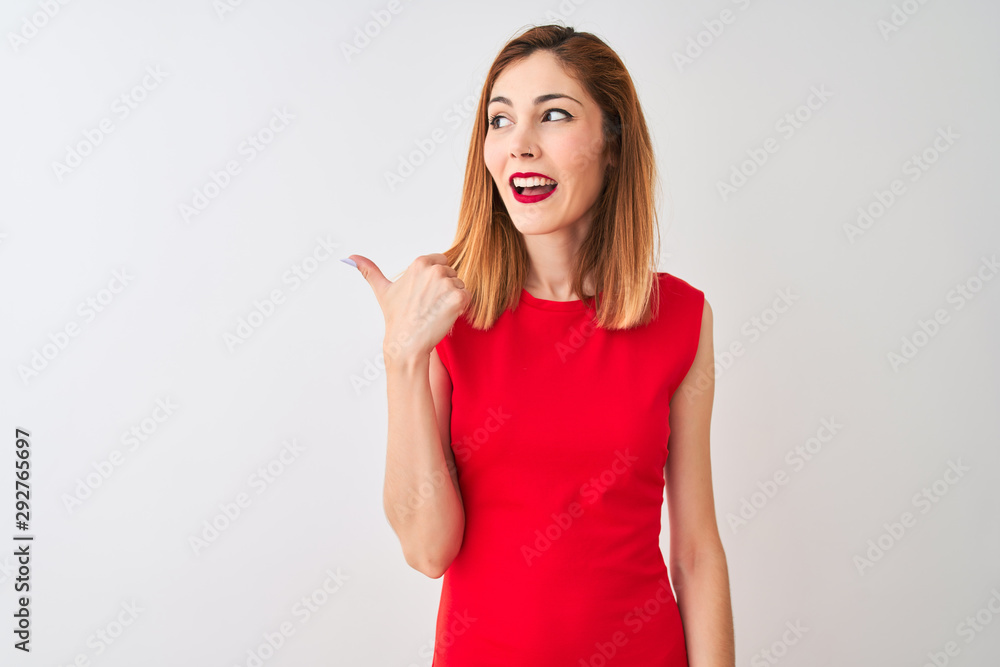 Redhead businesswoman wearing elegant red dress standing over isolated white background smiling with happy face looking and pointing to the side with thumb up. Redhead businesswoman wearing elegant red dress standing over isolated white background smiling with happy face looking and pointing to the side with thumb up.