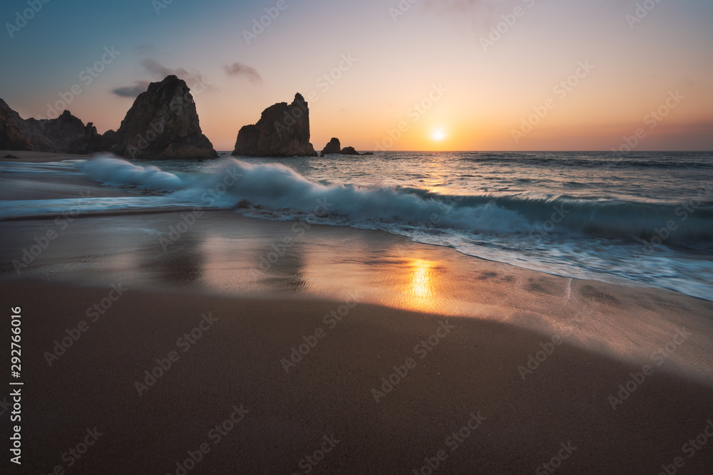 Portugal Ursa Beach at atlantic coast. Sunset reflection. White wave roll towards sandy beach with rock silhouette in background. Picturesque landscape panorama