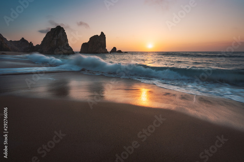 Portugal Ursa Beach at atlantic coast. Sunset reflection. White wave roll towards sandy beach with rock silhouette in background. Picturesque landscape panorama