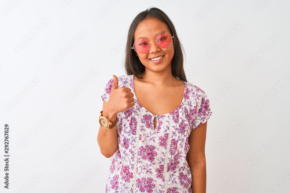Young chinese woman wearing floral t-shirt and pink glasses over isolated white background doing happy thumbs up gesture with hand. Approving expression looking at the camera with showing success. Young chinese woman wearing floral t-shirt and pink glasses over isolated white background doing happy thumbs up gesture with hand. Approving expression looking at the camera with showing success.