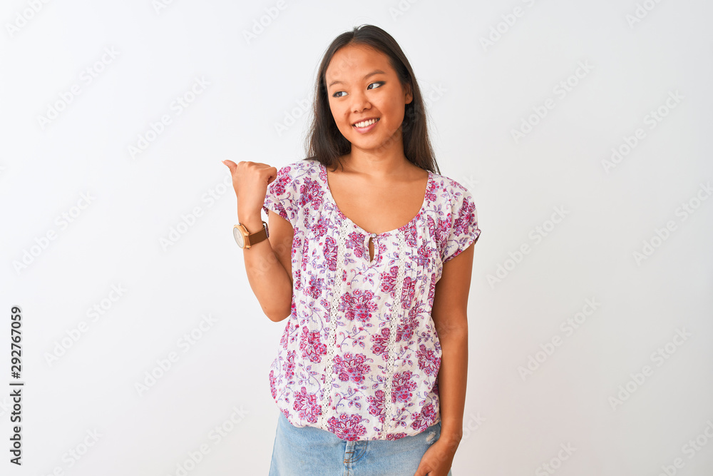 Young chinese woman wearing casual floral t-shirt standing over isolated white background smiling with happy face looking and pointing to the side with thumb up. Young chinese woman wearing casual floral t-shirt standing over isolated white background smiling with happy face looking and pointing to the side with thumb up.