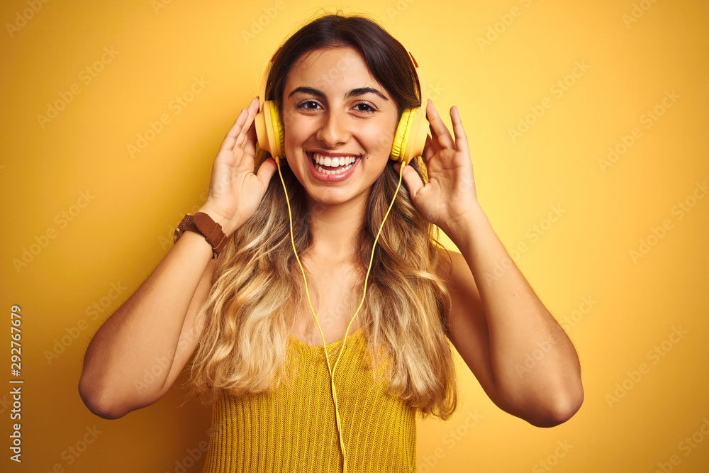 Young beautiful woman wearing headphones over yellow isolated background with a happy face standing and smiling with a confident smile showing teeth Young beautiful woman wearing headphones over yellow isolated background with a happy face standing and smiling with a confident smile showing teeth