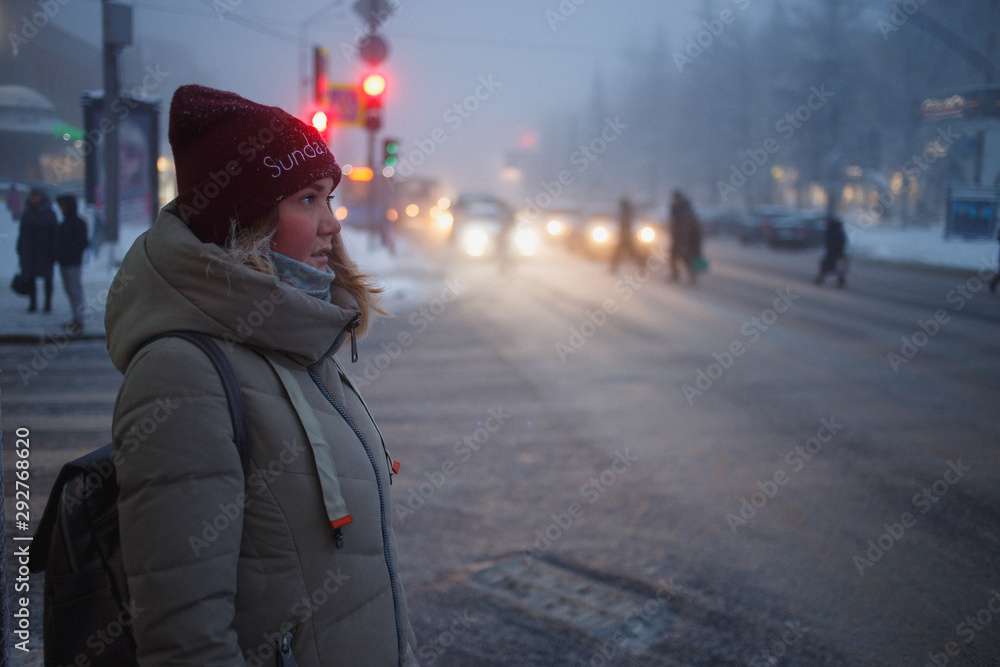 Woman walking in the city in the winter dusk time. The woman is waiting for a green traffic light signal on the crossroads. Woman walking in the city in the winter dusk time. The woman is waiting for a green traffic light signal on the crossroads.