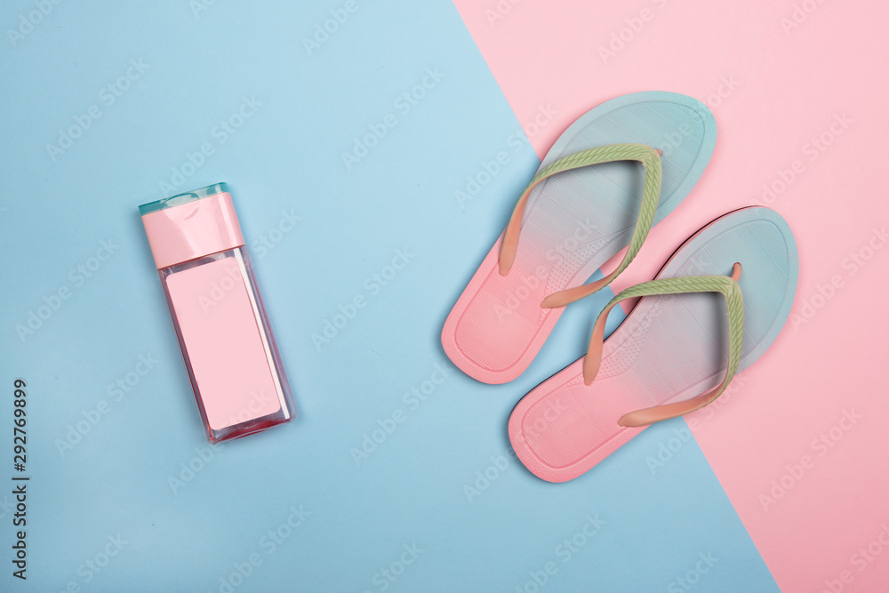 Stylish beach flip-flops and pink sunscreen bottle on pink and blue pastel background, top view. Summer minimalistic concept with copy space. Stylish beach flip-flops and pink sunscreen bottle on pink and blue pastel background, top view. Summer minimalistic concept with copy space.