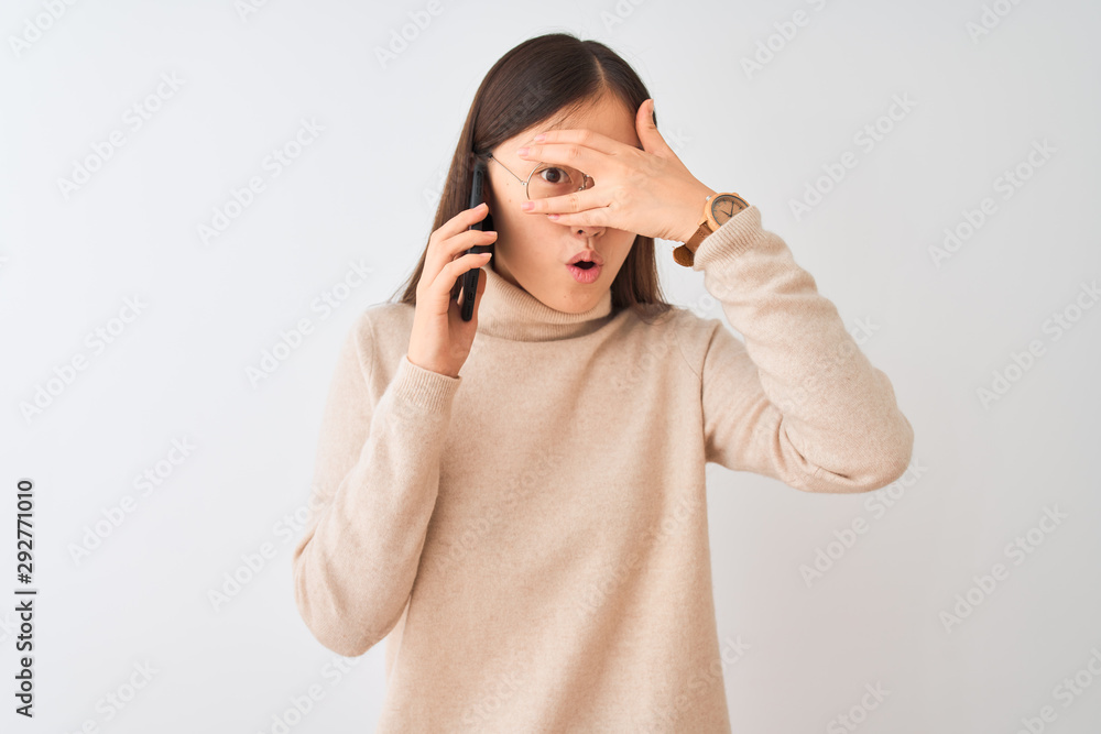 Young chinese woman talking on the smartphone over isolated white background peeking in shock covering face and eyes with hand, looking through fingers with embarrassed expression. Young chinese woman talking on the smartphone over isolated white background peeking in shock covering face and eyes with hand, looking through fingers with embarrassed expression.