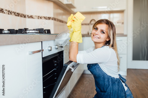 A young woman from a professional cleaning company cleans up at home. She is tin he kitchen in yellow gloves with cleaning supplies stuff near oven.
