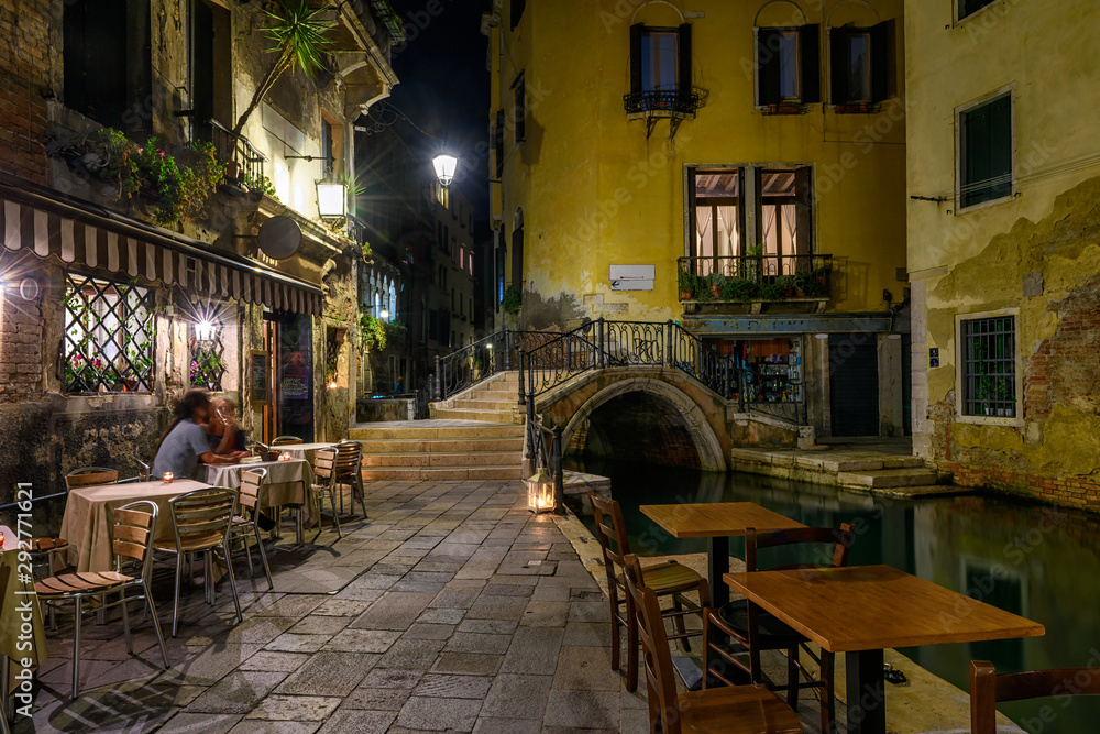 Narrow canal with bridge and tables of restaurant in Venice, Italy. Architecture and landmark of Venice. Night cozy cityscape of Venice.