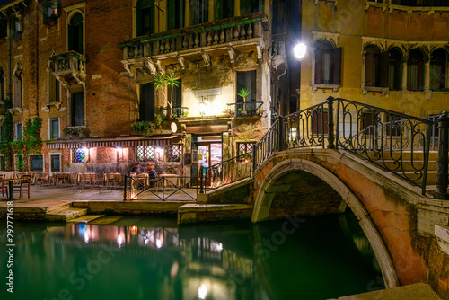Fototapeta Naklejka Na Ścianę i Meble -  Narrow canal with bridge and tables of restaurant in Venice, Italy. Architecture and landmark of Venice. Night cozy cityscape of Venice.