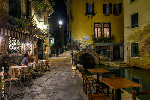 Narrow canal with bridge and tables of restaurant in Venice, Italy. Architecture and landmark of Venice. Night cozy cityscape of Venice.