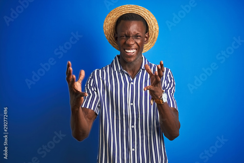 African american man wearing striped shirt and summer hat over isolated blue background celebrating mad and crazy for success with arms raised and closed eyes screaming excited. Winner concept