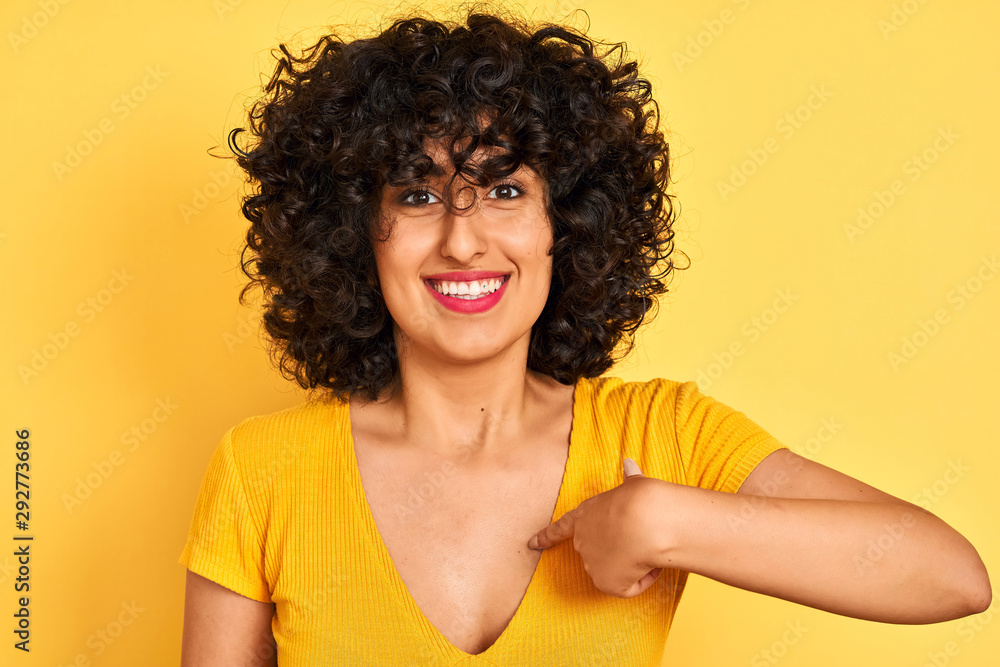 Young arab woman with curly hair wearing t-shirt standing over isolated yellow background with surprise face pointing finger to himself Young arab woman with curly hair wearing t-shirt standing over isolated yellow background with surprise face pointing finger to himself
