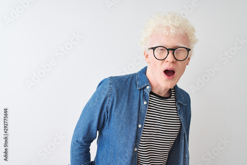 Young albino blond man wearing denim shirt and glasses over isolated white background In shock face, looking skeptical and sarcastic, surprised with open mouth