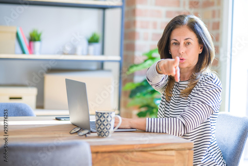 Middle age senior woman sitting at the table at home working using computer laptop pointing with finger to the camera and to you, hand sign, positive and confident gesture from the front