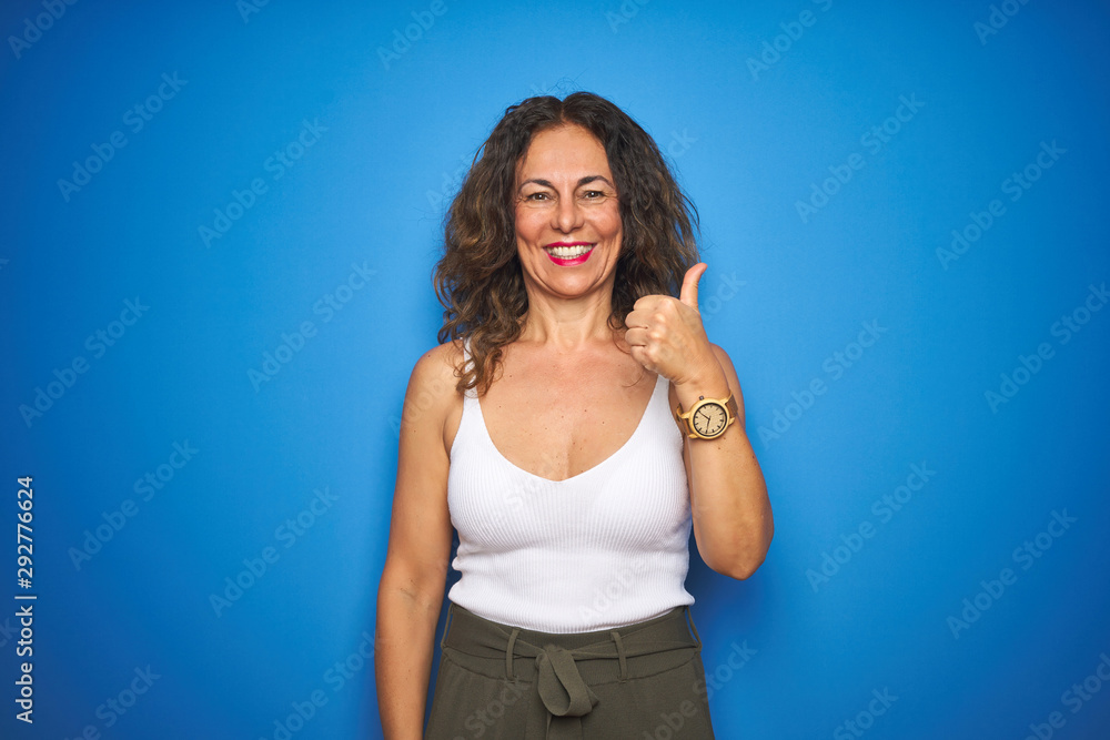 Middle age senior woman with curly hair standing over blue isolated background doing happy thumbs up gesture with hand. Approving expression looking at the camera showing success. Middle age senior woman with curly hair standing over blue isolated background doing happy thumbs up gesture with hand. Approving expression looking at the camera showing success.