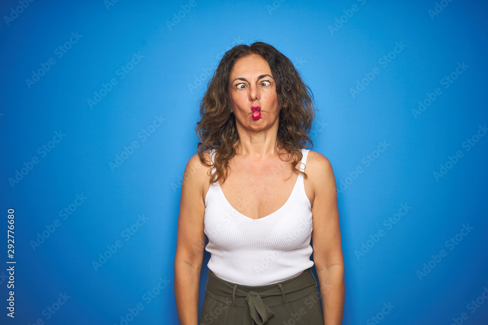 Middle age senior woman with curly hair standing over blue isolated background making fish face with lips, crazy and comical gesture. Funny expression. Middle age senior woman with curly hair standing over blue isolated background making fish face with lips, crazy and comical gesture. Funny expression.