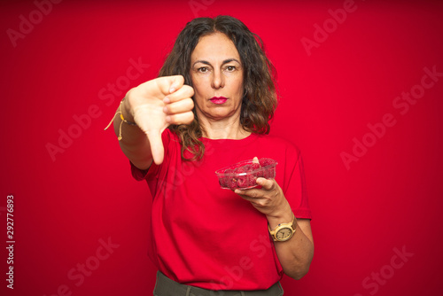 Middle age senior woman eating raspberries over red isolated background with angry face, negative sign showing dislike with thumbs down, rejection concept