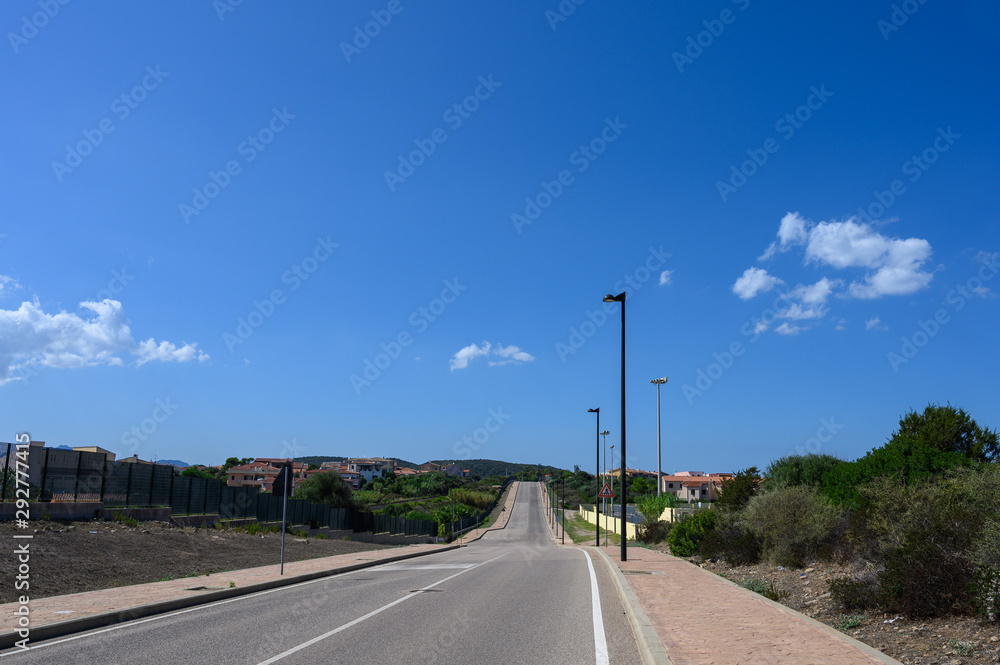 Road goes into the distance against a blue sky with clouds. On the sides trees and a small village Road goes into the distance against a blue sky with clouds. On the sides trees and a small village