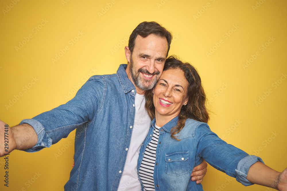 Naklejka premium Beautiful middle age couple together wearing denim shirt over isolated yellow background looking at the camera smiling with open arms for hug. Cheerful expression embracing happiness.