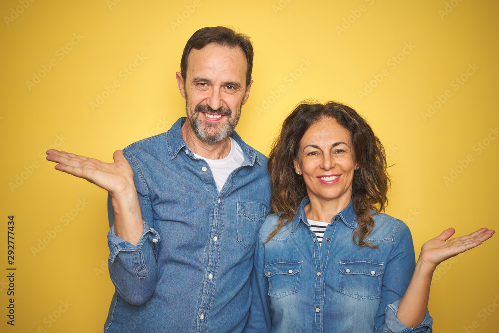 Beautiful middle age couple together standing over isolated yellow background smiling cheerful presenting and pointing with palm of hand looking at the camera. Beautiful middle age couple together standing over isolated yellow background smiling cheerful presenting and pointing with palm of hand looking at the camera.