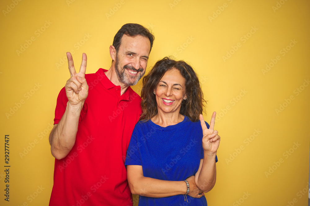 Beautiful middle age couple together standing over isolated yellow background smiling with happy face winking at the camera doing victory sign. Number two. Beautiful middle age couple together standing over isolated yellow background smiling with happy face winking at the camera doing victory sign. Number two.