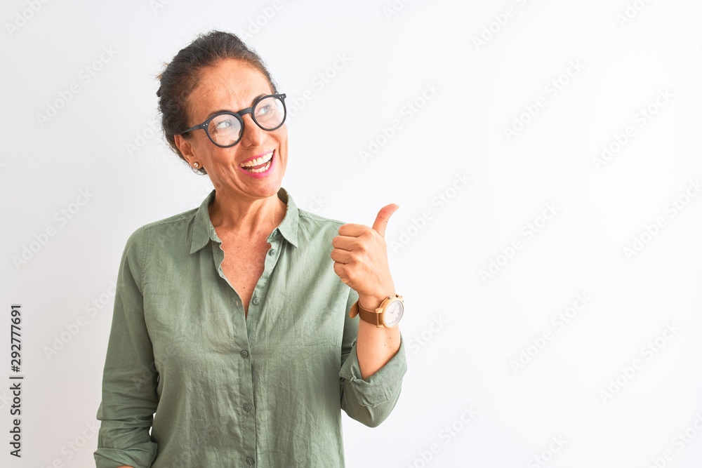 Middle age woman wearing green shirt and glasses standing over isolated white background smiling with happy face looking and pointing to the side with thumb up. Middle age woman wearing green shirt and glasses standing over isolated white background smiling with happy face looking and pointing to the side with thumb up.