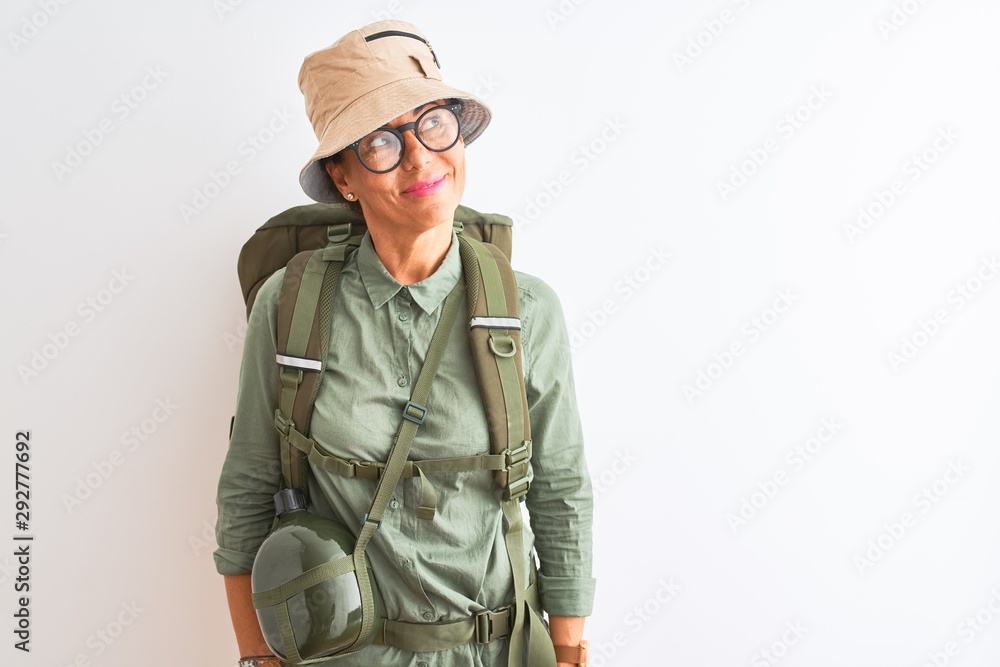Middle age hiker woman wearing backpack canteen hat glasses over isolated white background smiling looking to the side and staring away thinking. Middle age hiker woman wearing backpack canteen hat glasses over isolated white background smiling looking to the side and staring away thinking.