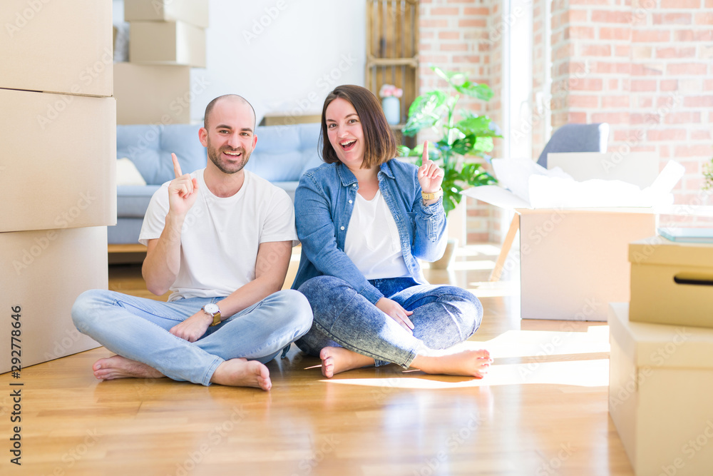 Young couple sitting on the floor arround cardboard boxes moving to a new house pointing finger up with successful idea. Exited and happy. Number one. Young couple sitting on the floor arround cardboard boxes moving to a new house pointing finger up with successful idea. Exited and happy. Number one.