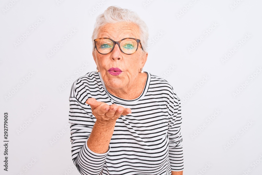 Senior grey-haired woman wearing striped navy t-shirt glasses over isolated white background looking at the camera blowing a kiss with hand on air being lovely and sexy. Love expression. Senior grey-haired woman wearing striped navy t-shirt glasses over isolated white background looking at the camera blowing a kiss with hand on air being lovely and sexy. Love expression.