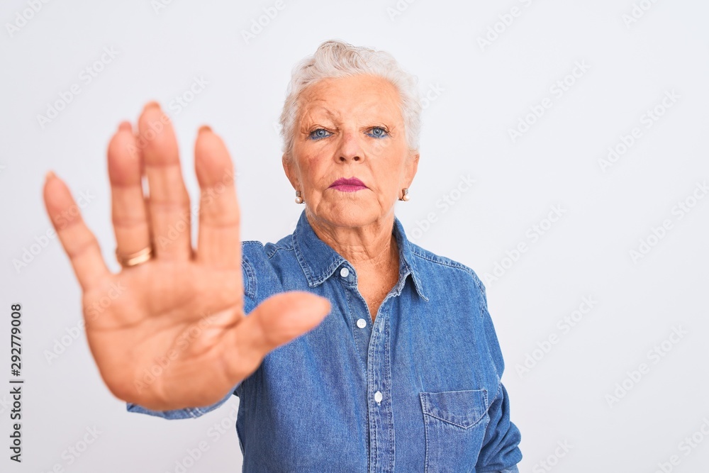 Senior grey-haired woman wearing casual denim shirt standing over isolated white background with open hand doing stop sign with serious and confident expression, defense gesture Senior grey-haired woman wearing casual denim shirt standing over isolated white background with open hand doing stop sign with serious and confident expression, defense gesture