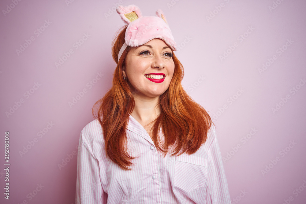 Young beautiful redhead woman wearing pajama over pink isolated background looking away to side with smile on face, natural expression. Laughing confident. Young beautiful redhead woman wearing pajama over pink isolated background looking away to side with smile on face, natural expression. Laughing confident.