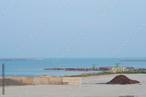 A pile of wood chips is resting beside concrete blocks and gravel on an industrial port. The port is closed. The sky is clear, and the water calm. A concrete breakwater is near the port.