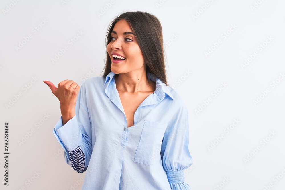 Young beautiful woman wearing blue elegant shirt standing over isolated white background smiling with happy face looking and pointing to the side with thumb up. Young beautiful woman wearing blue elegant shirt standing over isolated white background smiling with happy face looking and pointing to the side with thumb up.