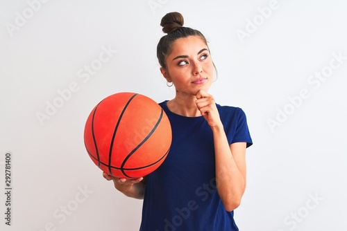 Young beautiful sportswoman holding basketball ball over isolated white background serious face thinking about question, very confused idea © Krakenimages.com