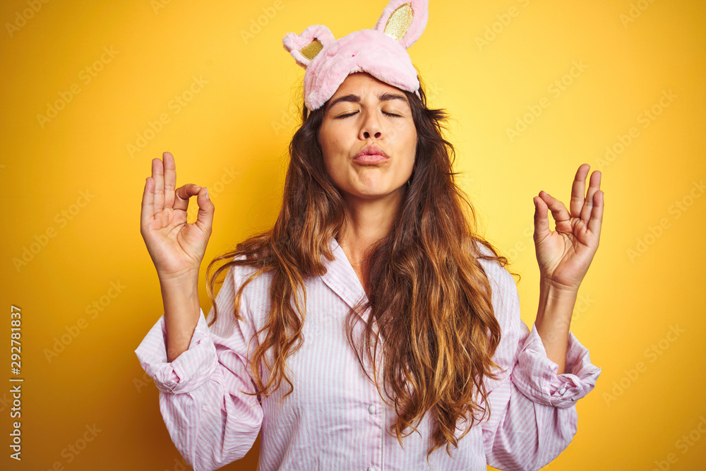 Young woman wearing pajama and sleep mask standing over yellow isolated background relaxed and smiling with eyes closed doing meditation gesture with fingers. Yoga concept. Young woman wearing pajama and sleep mask standing over yellow isolated background relaxed and smiling with eyes closed doing meditation gesture with fingers. Yoga concept.