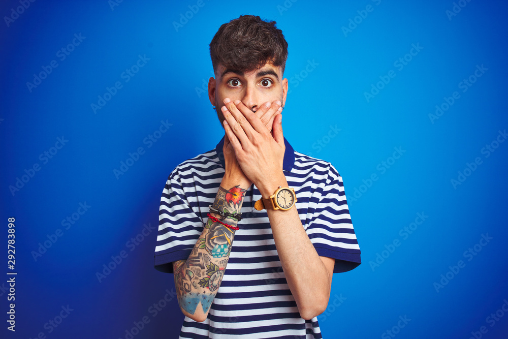 Young man with tattoo wearing striped polo standing over isolated blue background shocked covering mouth with hands for mistake. Secret concept. Young man with tattoo wearing striped polo standing over isolated blue background shocked covering mouth with hands for mistake. Secret concept.