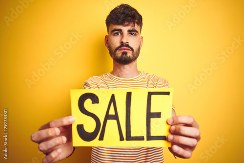 Young man with tattoo holding sale banner standing over isolated yellow background with a confident expression on smart face thinking serious