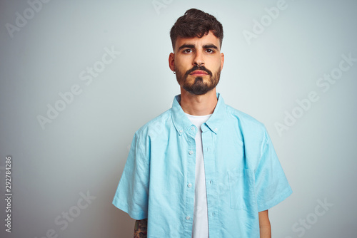 Young man with tattoo wearing blue shirt standing over isolated white background with serious expression on face. Simple and natural looking at the camera.