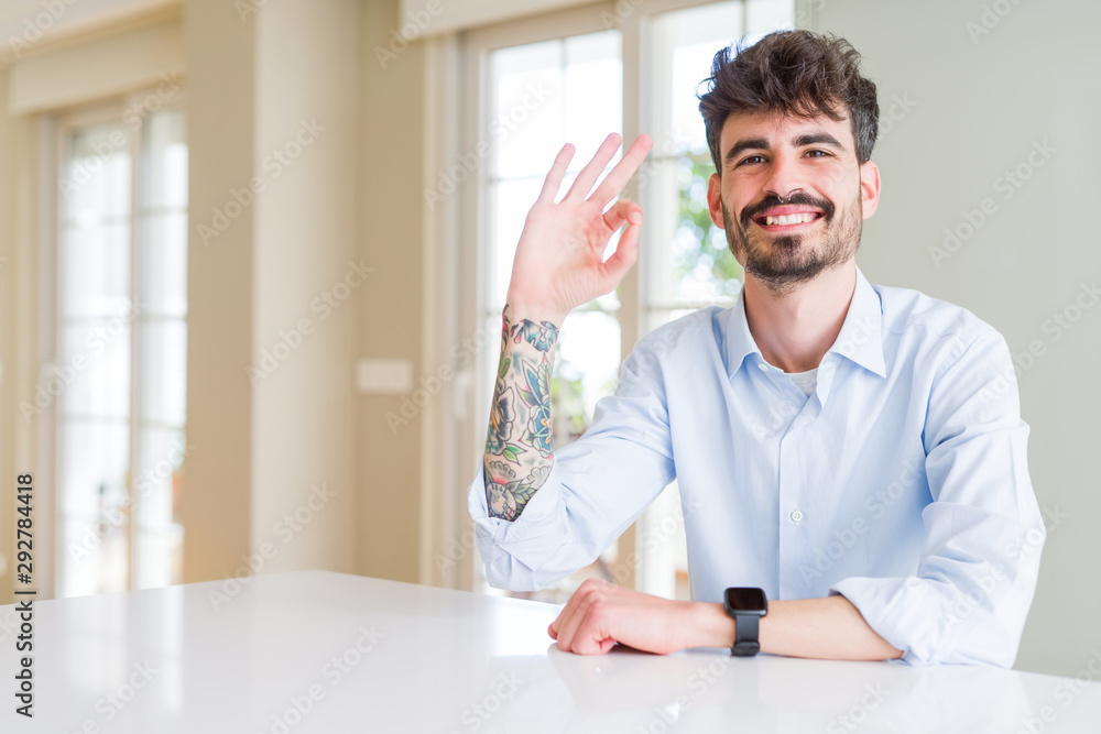 Young businesss man sitting on white table smiling positive doing ok sign with hand and fingers. Successful expression. Young businesss man sitting on white table smiling positive doing ok sign with hand and fingers. Successful expression.