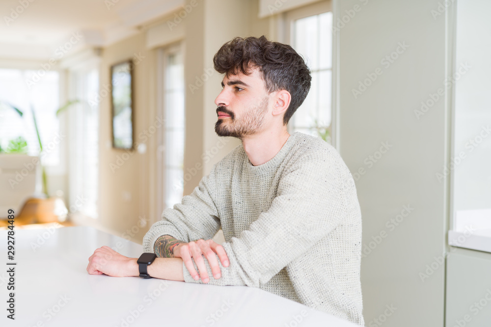Young man wearing casual sweater sitting on white table looking to side, relax profile pose with natural face with confident smile.