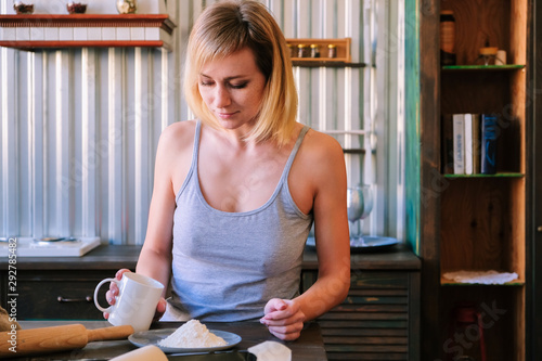 A beautiful freckled blonde girl in pajamas is standing near the kitchen table, drinking Fock, tea and preparing pastries.