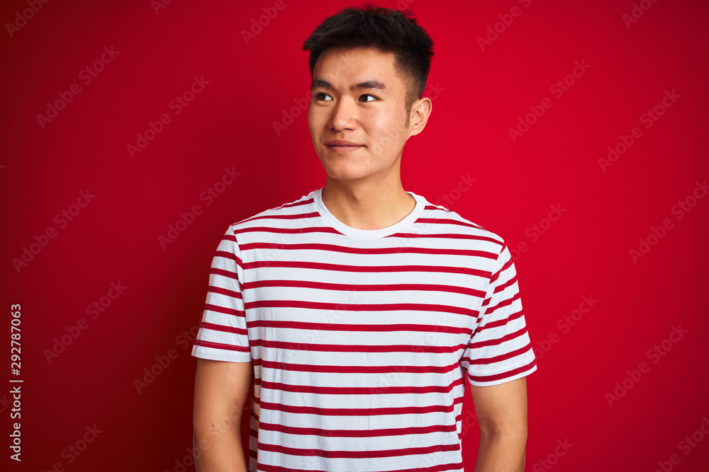 Young asian chinese man wearing striped t-shirt standing over isolated red background smiling looking to the side and staring away thinking. Young asian chinese man wearing striped t-shirt standing over isolated red background smiling looking to the side and staring away thinking.