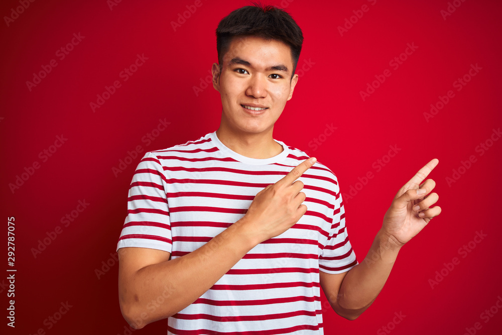 Young asian chinese man wearing striped t-shirt standing over isolated red background smiling and looking at the camera pointing with two hands and fingers to the side. Young asian chinese man wearing striped t-shirt standing over isolated red background smiling and looking at the camera pointing with two hands and fingers to the side.