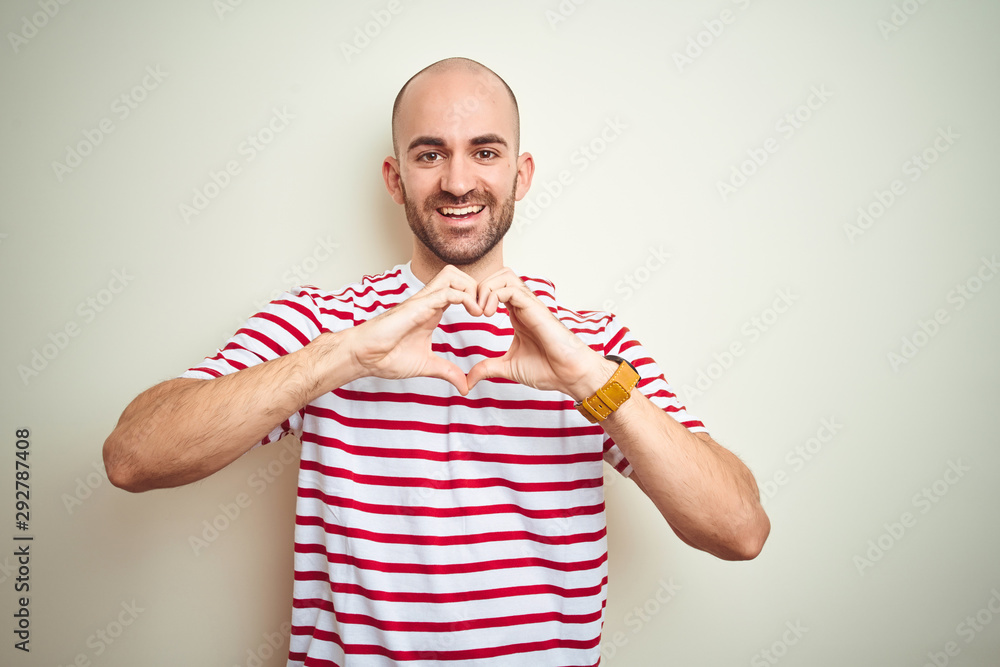 Naklejka premium Young bald man with beard wearing casual striped red t-shirt over white isolated background smiling in love doing heart symbol shape with hands. Romantic concept.