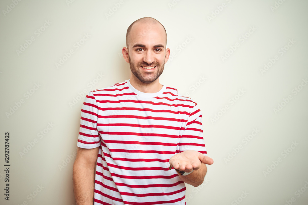 Young bald man with beard wearing casual striped red t-shirt over white isolated background smiling cheerful offering palm hand giving assistance and acceptance. Young bald man with beard wearing casual striped red t-shirt over white isolated background smiling cheerful offering palm hand giving assistance and acceptance.