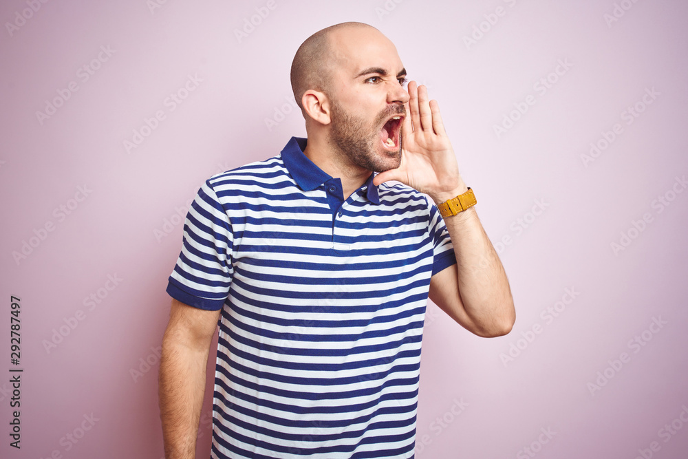 Young bald man with beard wearing casual striped blue t-shirt over pink isolated background shouting and screaming loud to side with hand on mouth. Communication concept. Young bald man with beard wearing casual striped blue t-shirt over pink isolated background shouting and screaming loud to side with hand on mouth. Communication concept.