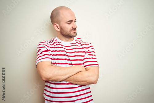Young bald man with beard wearing casual striped red t-shirt over white isolated background looking to the side with arms crossed convinced and confident