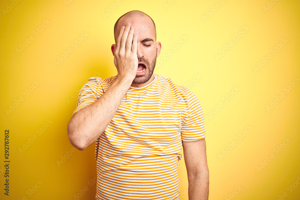 Young bald man with beard wearing casual striped t-shirt over yellow isolated background Yawning tired covering half face, eye and mouth with hand. Face hurts in pain. Young bald man with beard wearing casual striped t-shirt over yellow isolated background Yawning tired covering half face, eye and mouth with hand. Face hurts in pain.