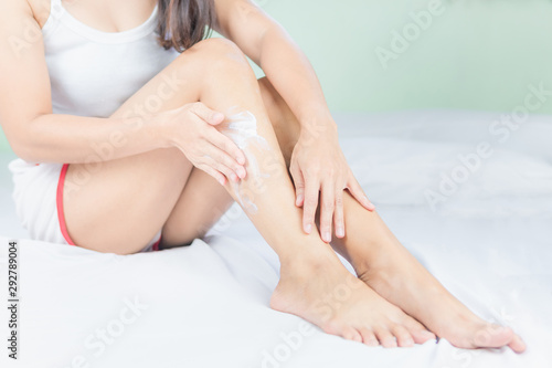 Beautiful young asian woman sitting on a bed stroking legs with soft smooth skin in the bedroom  girl applying touch body cream and lotion with treatment care concept.