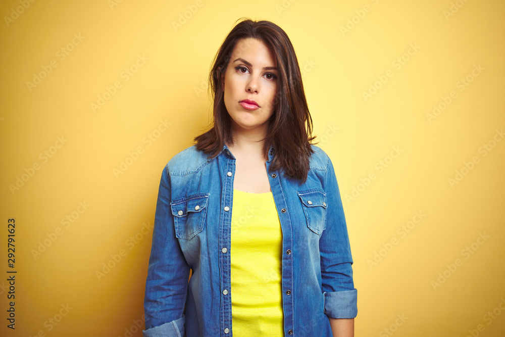 Young beautiful brunette woman wearing casual denim jacket over yellow isolated background with serious expression on face. Simple and natural looking at the camera. Young beautiful brunette woman wearing casual denim jacket over yellow isolated background with serious expression on face. Simple and natural looking at the camera.