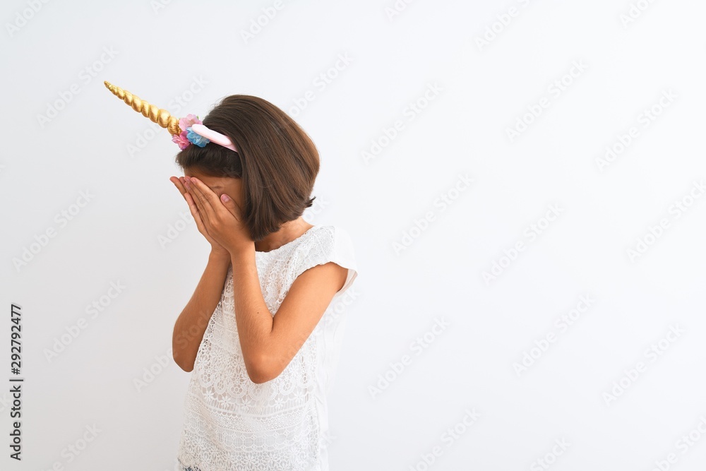 Beautiful child girl wearing unicorn diadem standing over isolated white background with sad expression covering face with hands while crying. Depression concept. Beautiful child girl wearing unicorn diadem standing over isolated white background with sad expression covering face with hands while crying. Depression concept.